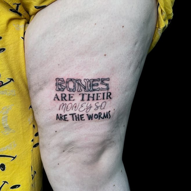 Tattoo on person's upper thigh reading "Bones are their money so are the worms" in stylized and handwritten font.