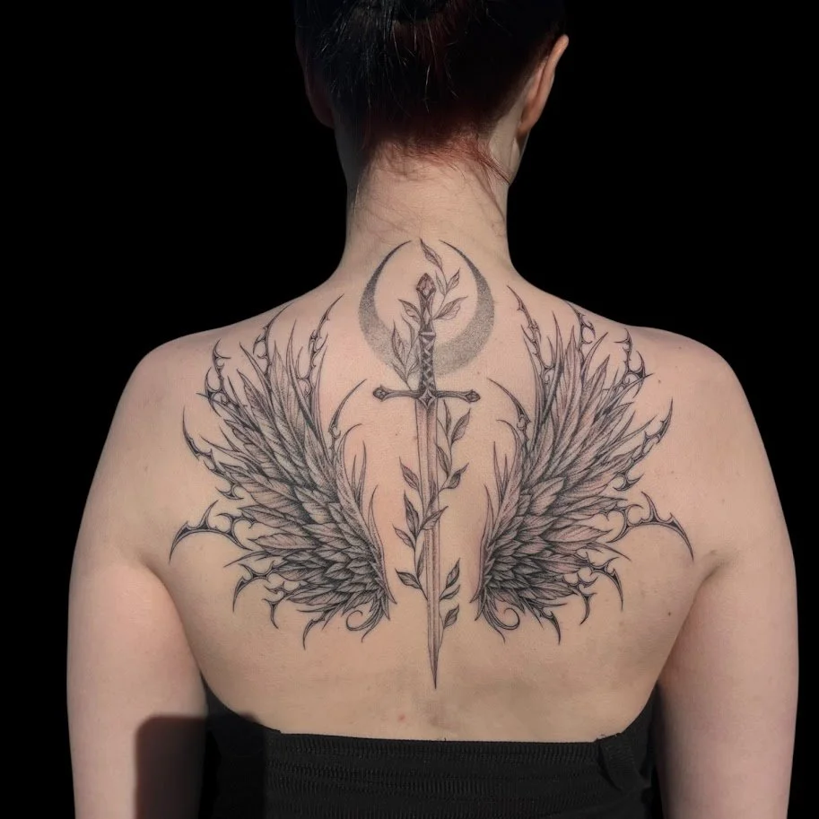Tattoo of a sword with wings on the person's upper back and a moon with a leaf on the neck.