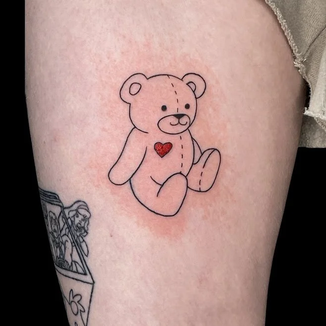 Close-up of a teddy bear tattoo with a red heart on its chest on a person's skin.