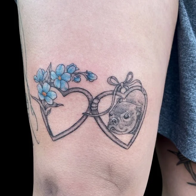 Tattoo of two overlapping hearts with blue flowers on one heart, and a dog inside the other heart, on a person's upper arm.