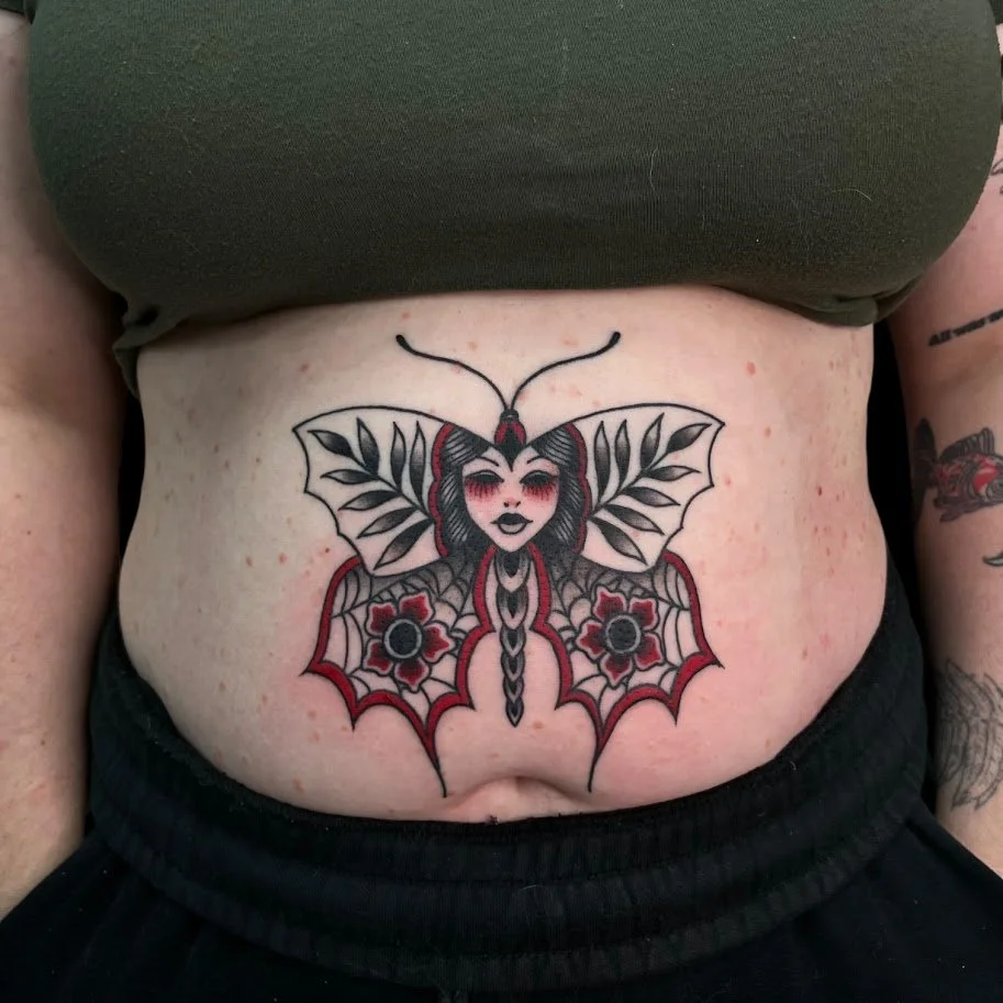 Tattoo of a butterfly with a woman's face, detailed wings, and floral motifs on the stomach.