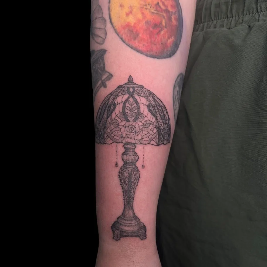 Line art tattoo of a vintage desk lamp on a person's forearm.