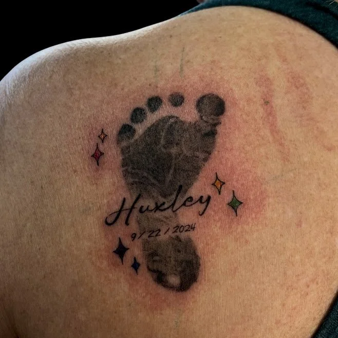 Tattoo of a baby footprint with the name 'Huxley' and the date '9/22/2024' surrounded by colorful star-like shapes.