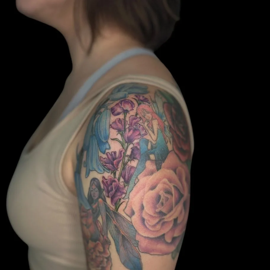 A close-up of a woman's shoulder tattoo featuring pink roses, purple flowers, blue leaves, and a small fairy with long dark hair.