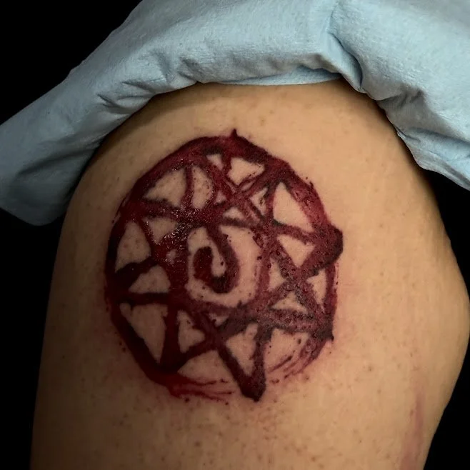 Fresh tattoo on skin featuring a dark red inked geometric design, including a central symbol surrounded by interconnected lines and shapes.