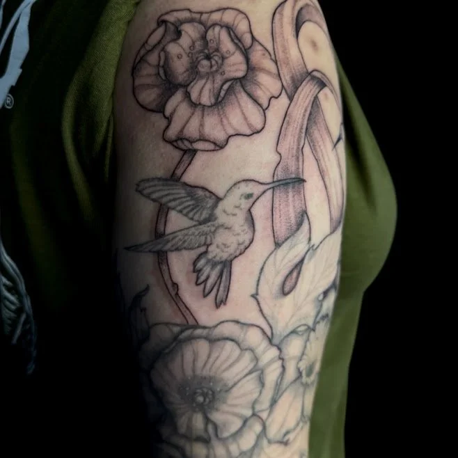 Black and gray tattoo of a hummingbird flying beside flowers and a snake on a person's arm.