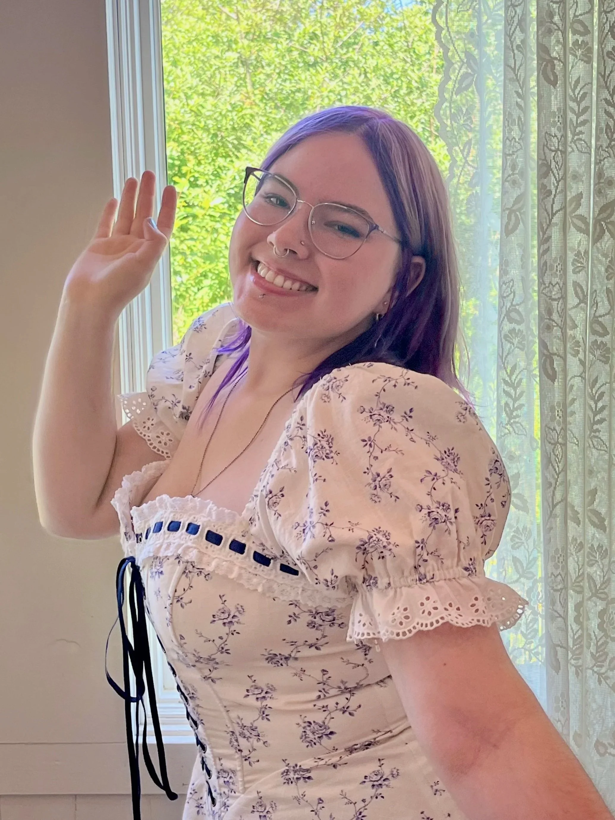 A young woman with glasses and purple hair smiling and waving, standing indoors near a window with lace curtains and green foliage outside.