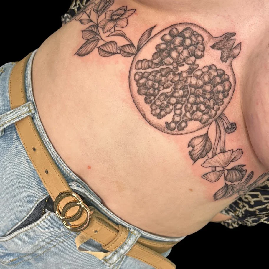 A black and gray tattoo of a pomegranate fruit with sections of seeds and leaves, located on the person's shoulder and upper chest area.