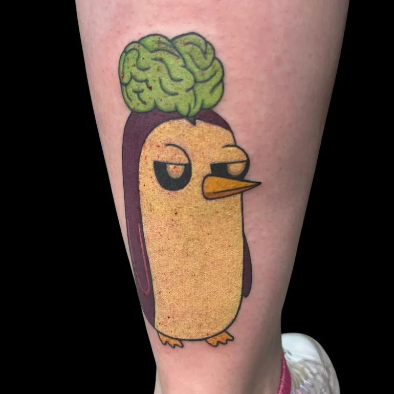 A tattoo of a cartoon penguin with a green brain on its head, a neutral expression, and orange beak, on a person's leg.
