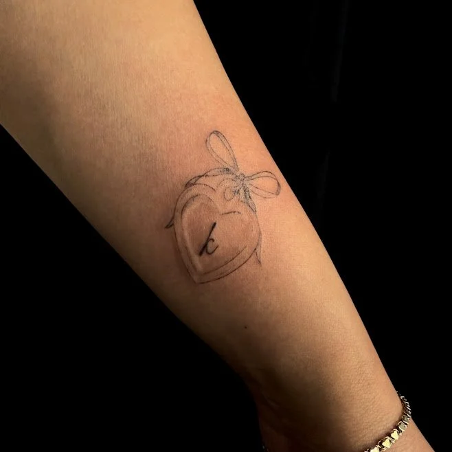 A tattoo of a simple outline of a bell with a bow on top, and the number 2 inside the bell, on someone's forearm.