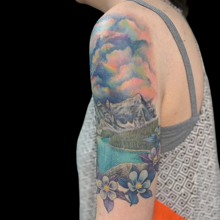 Colorful tattoo on a person's upper arm depicting clouds, mountains, a lake, and flowers with purple and yellow accents.