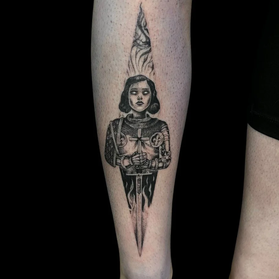 Black and gray tattoo of a woman with short hair, holding a sword vertically, with a flame-like element above her head.