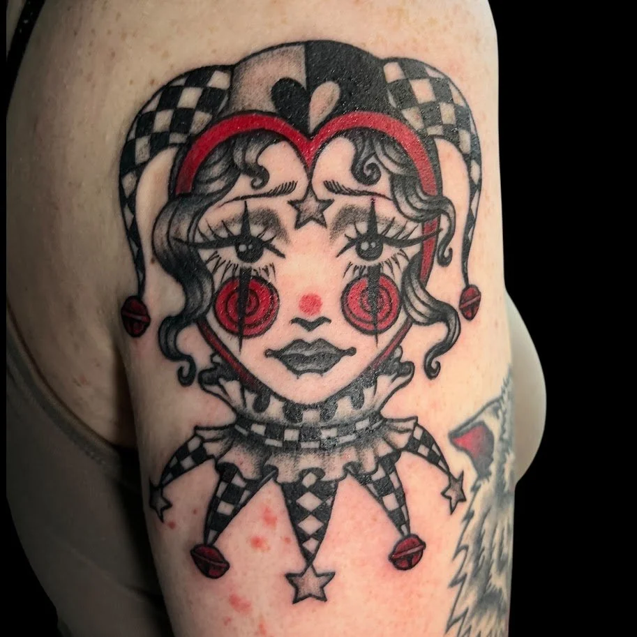 A colorful tattoo of a clown girl with a heart-shaped face, styled in a vintage circus theme, with checkered and red accents.