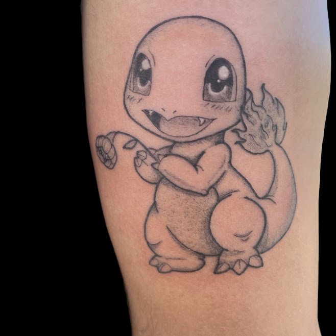 Black and gray tattoo of Charmander, a Pokémon character, on skin, showing Charmander with large eyes, a happy expression with an open mouth, holding a small item, and a flame on its tail.