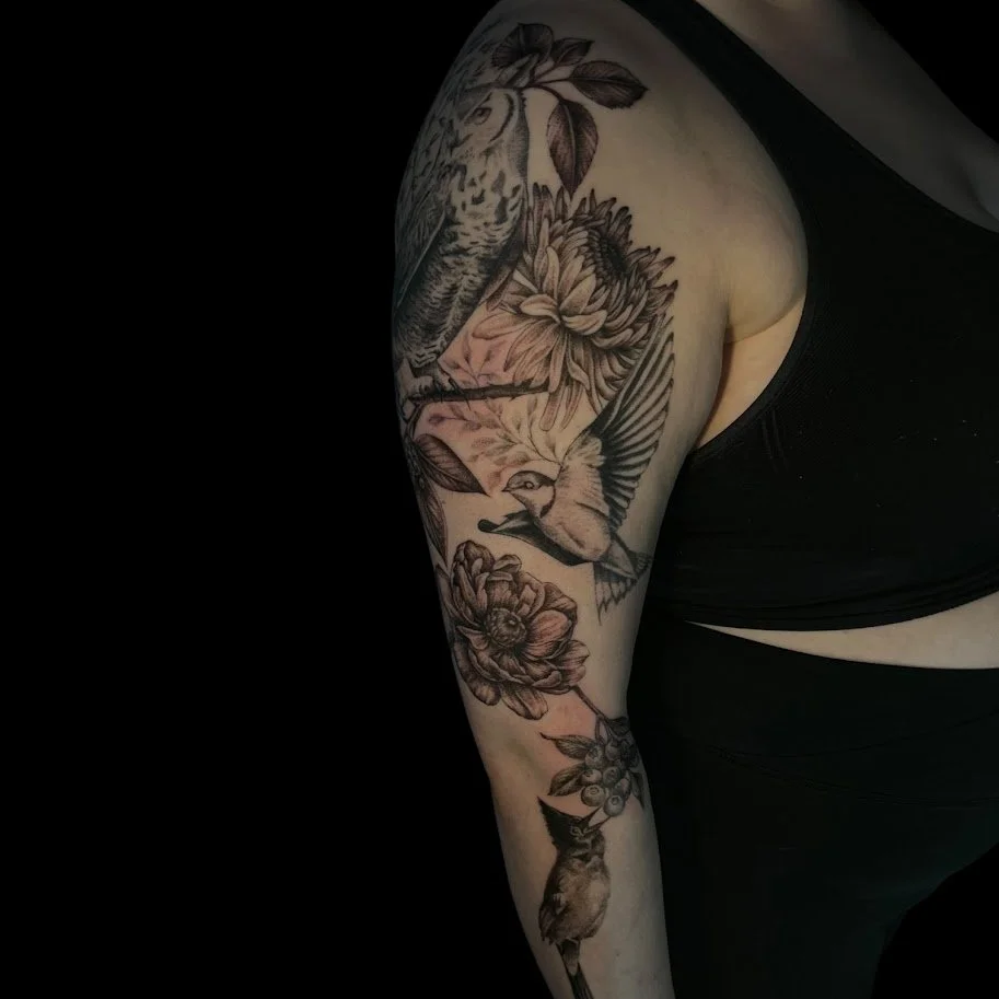 Black and gray tattoo sleeve featuring a owl, sunflower, hummingbird, rose, grapes, and a cat on a person's upper arm and shoulder.