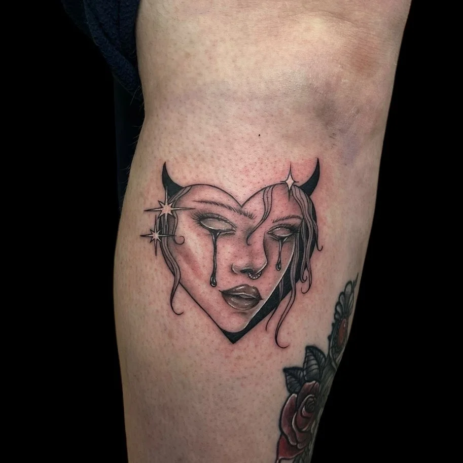 Tattoo of a woman's crying face with small horns and star charms, inside a heart shape.