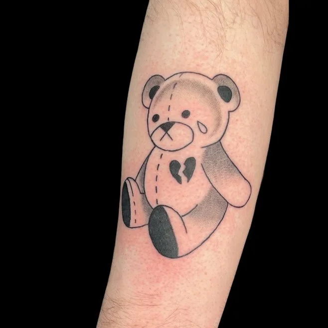 Tattoo of a sad teddy bear with a tear and a broken heart symbol on its chest.