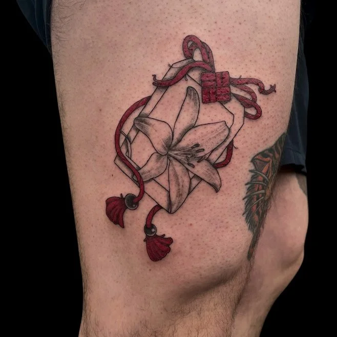 Tattoo of a white lily flower with red ribbon and bow on upper thigh.