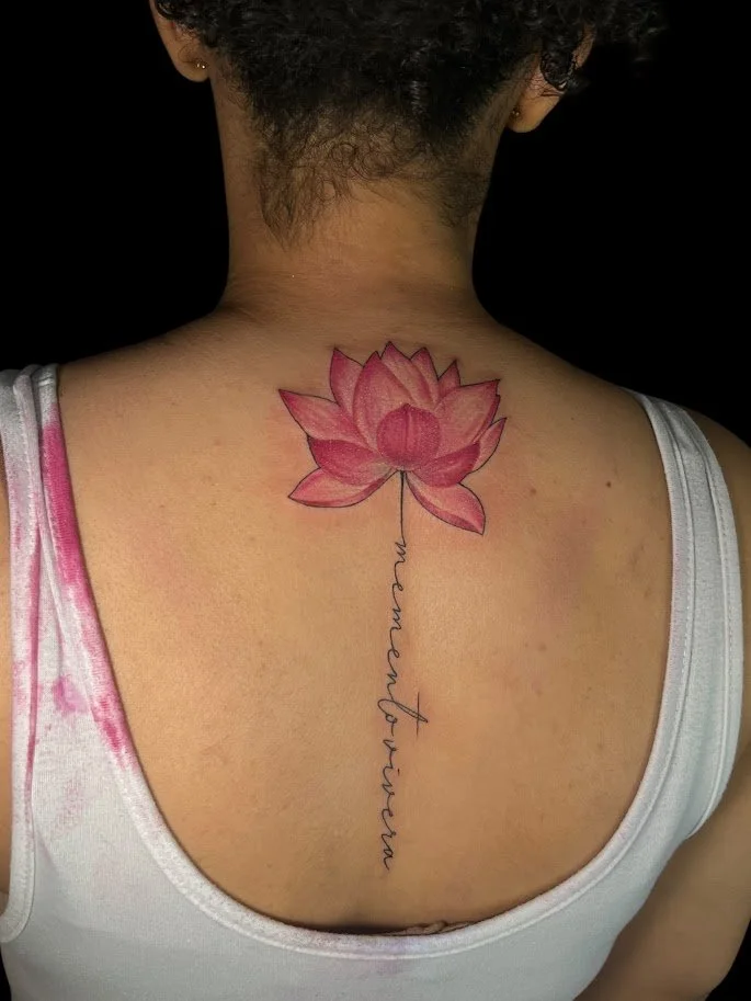Tattoo of a pink lotus flower on a person's upper back with cursive text hanging from the flower.
