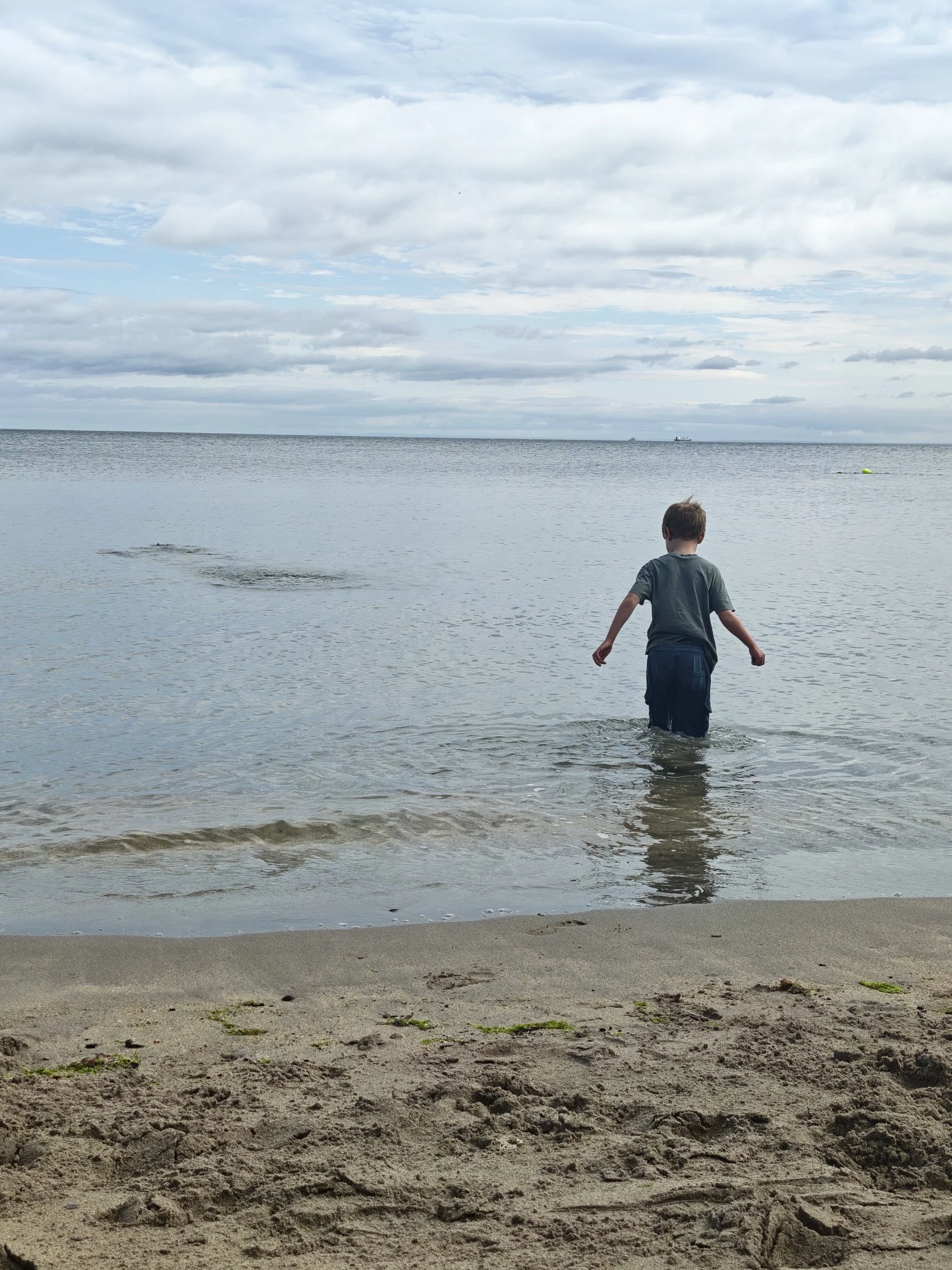 A young boy standing in shallow water at a beach with sandy shore, facing towards the ocean under a cloudy sky.