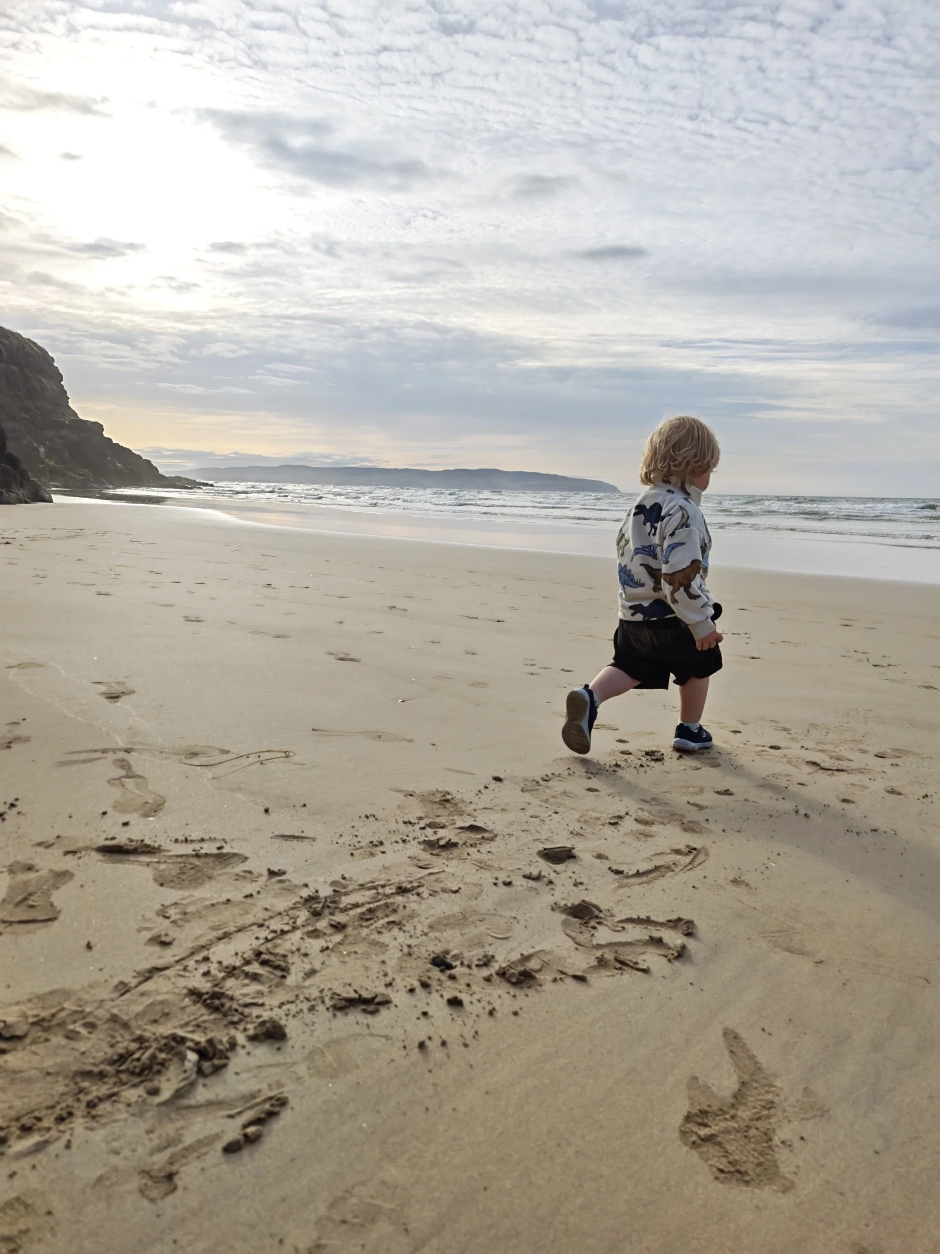 A young child with blonde hair, wearing a colorful dinosaur-patterned jacket, black shorts, and sneakers, walking along a sandy beach with footprints leading behind him. The beach is next to the ocean with waves crashing and a cliff on the left side. The sky is partly cloudy.