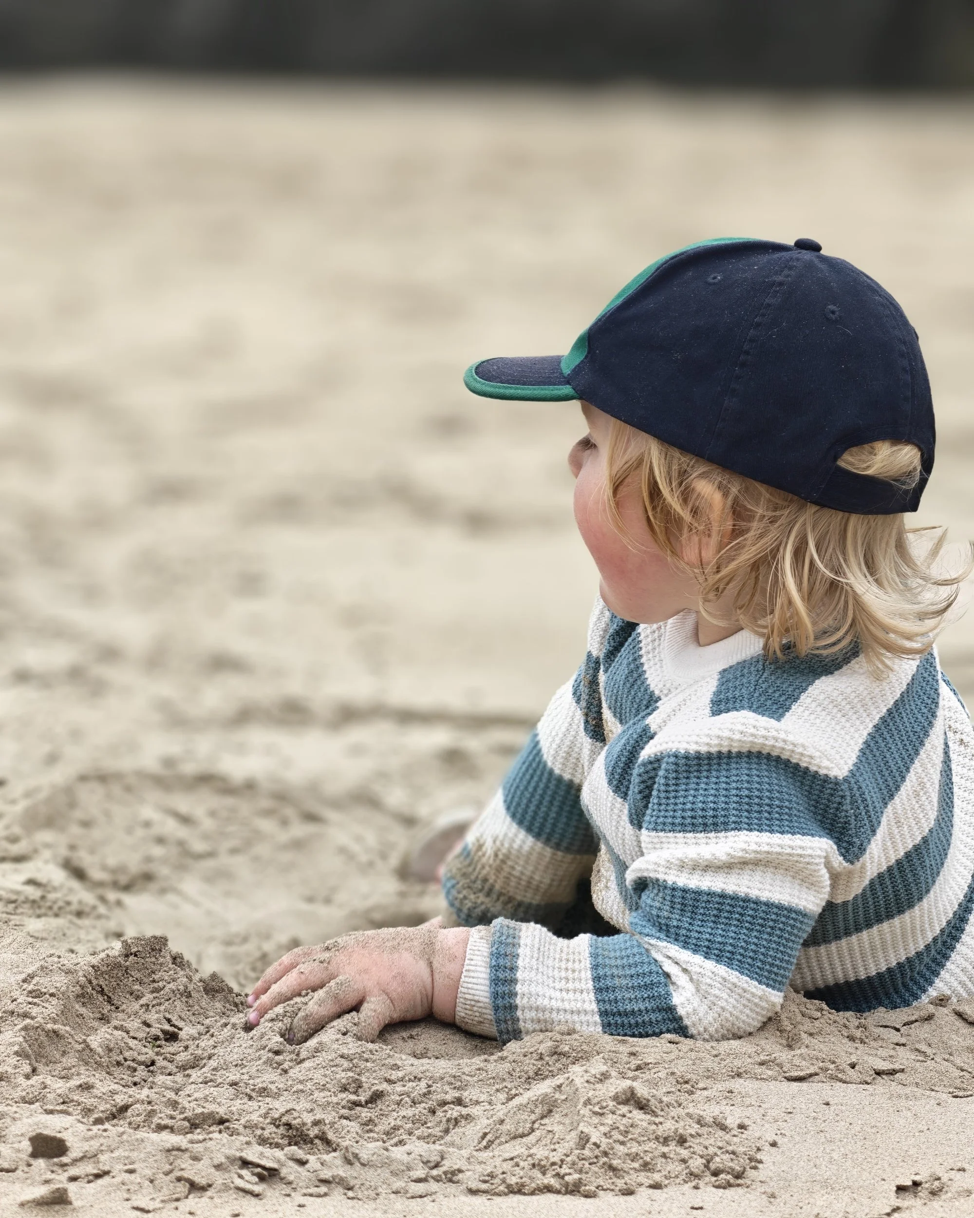 Child sitting on sandy beach, wearing a blue and white striped sweater and a navy blue cap, playing with the sand.