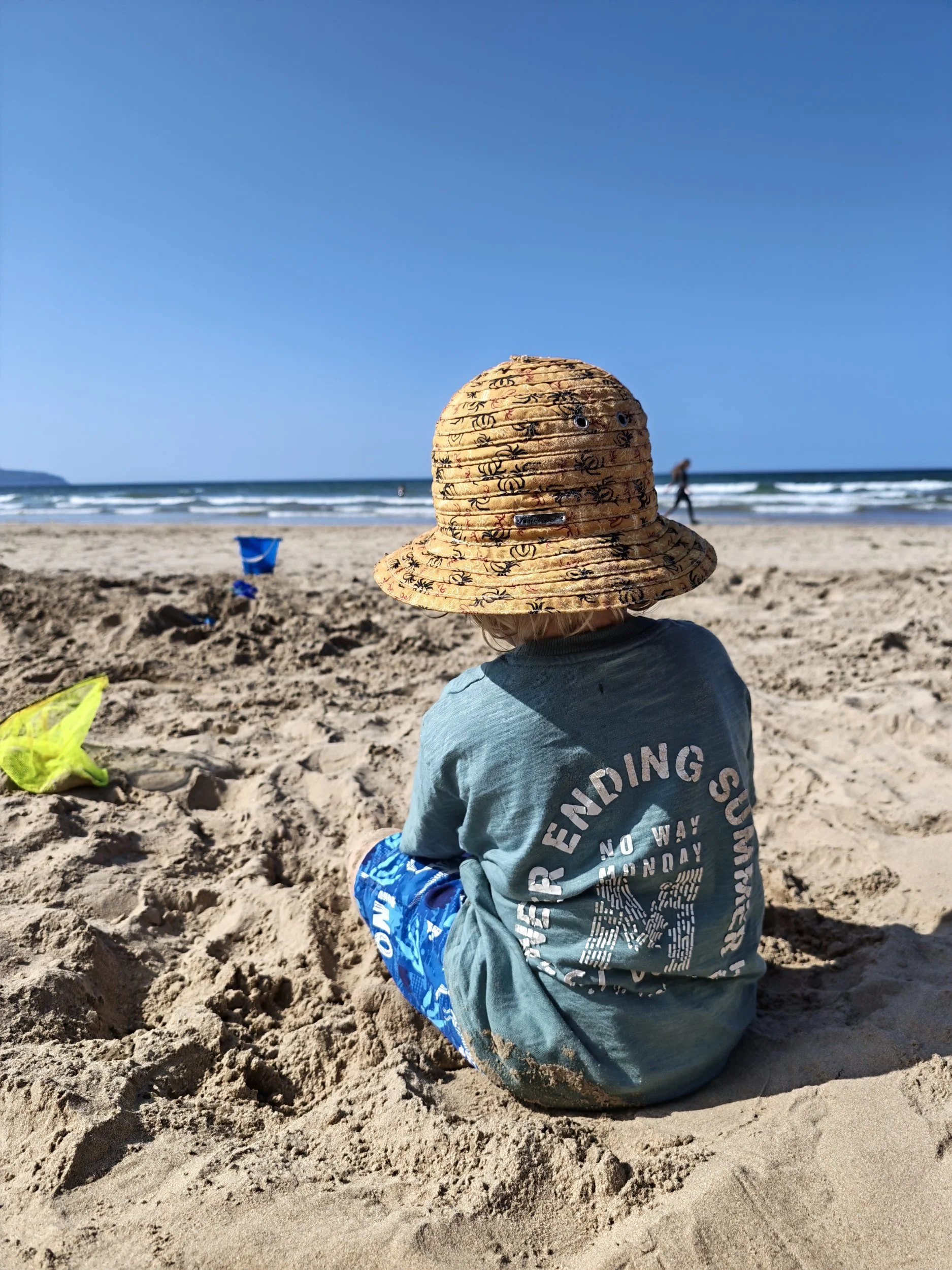 Child wearing a wide-brimmed straw hat and a gray T-shirt sitting on sandy beach, with ocean and waves in background and a person walking along the shoreline.