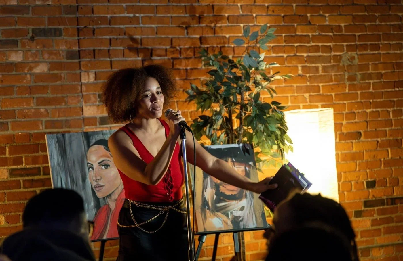 A woman with curly hair in a red top is speaking into a microphone in front of an audience. Behind her, there are two portraits and a potted plant, with a brick wall and a lamp in the background.