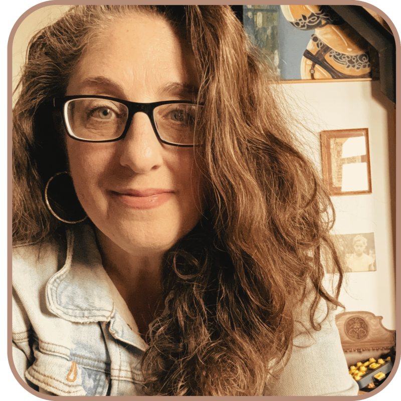 A woman with long, curly brown hair, glasses, and hoop earrings smiling for a selfie indoors.