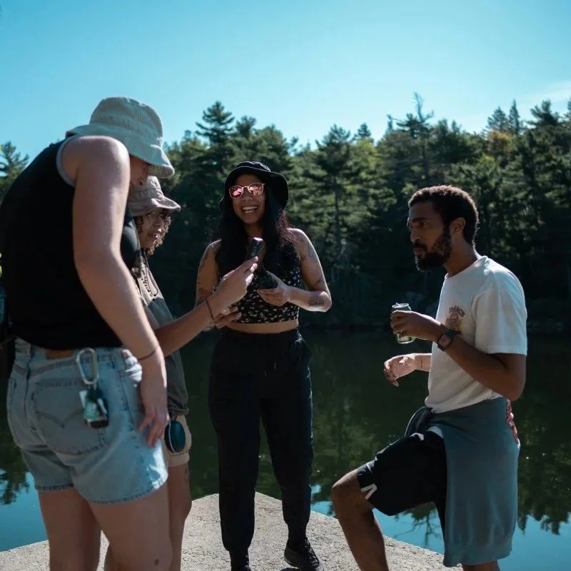Four young adults standing on a dock by a lake, smiling and looking at a phone, with trees in the background.