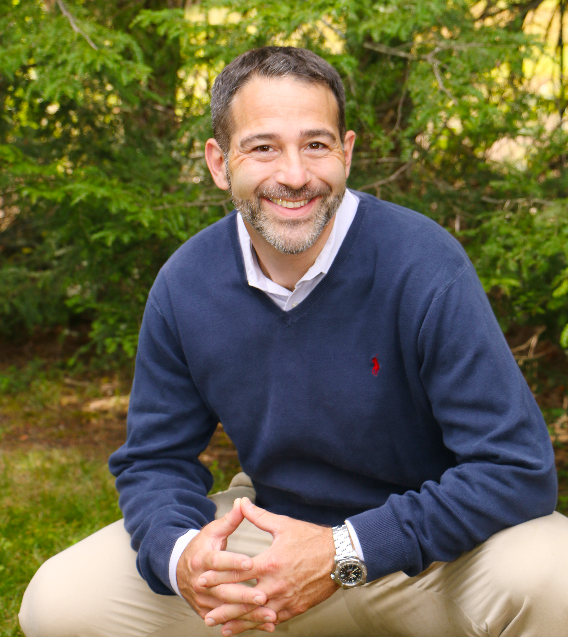 A smiling man with short dark hair, a beard, and wearing a navy blue sweater over a white shirt, sitting outdoors with green trees in the background, holding hands in a triangle shape in front of him.