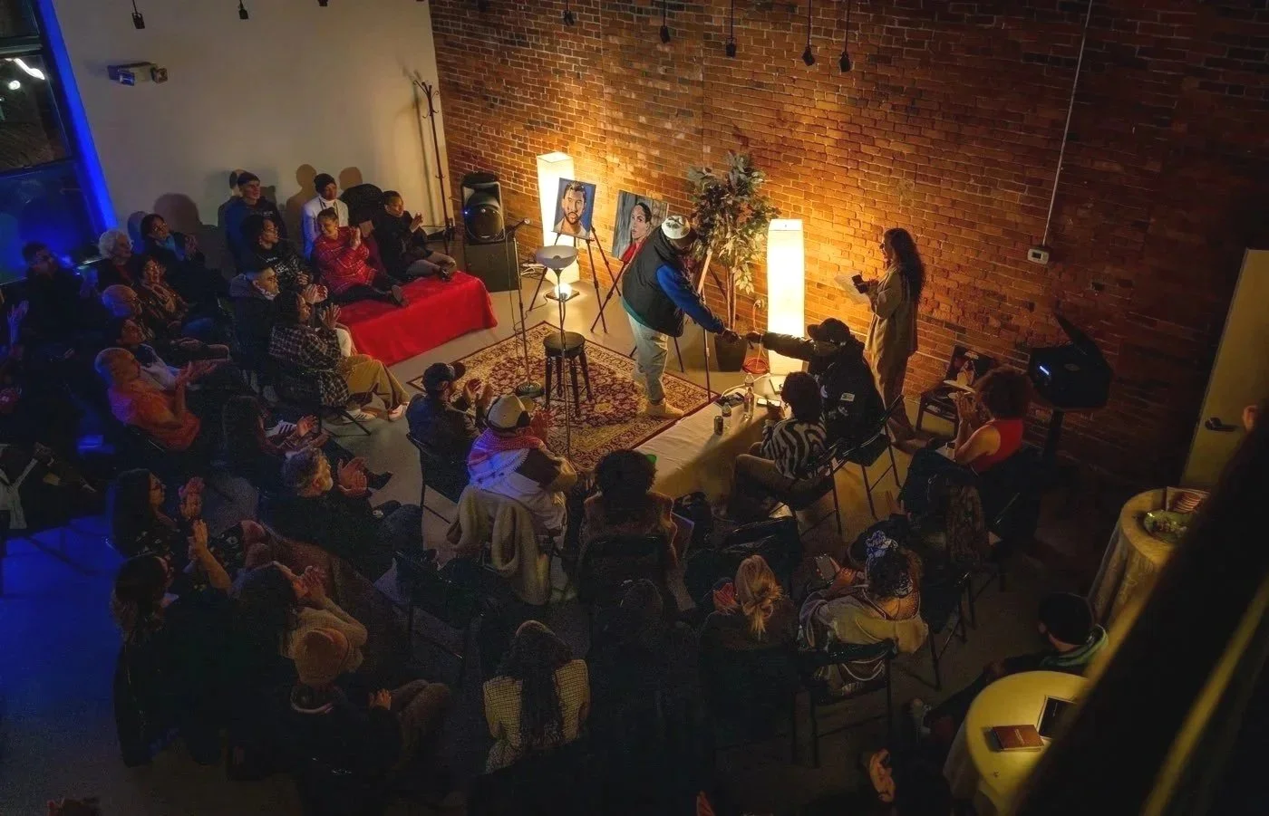 An art event or performance taking place in a cozy, dimly lit venue with an audience seated and clapping, two artists working on portraits, and paintings of people displayed on easels against a brick wall.