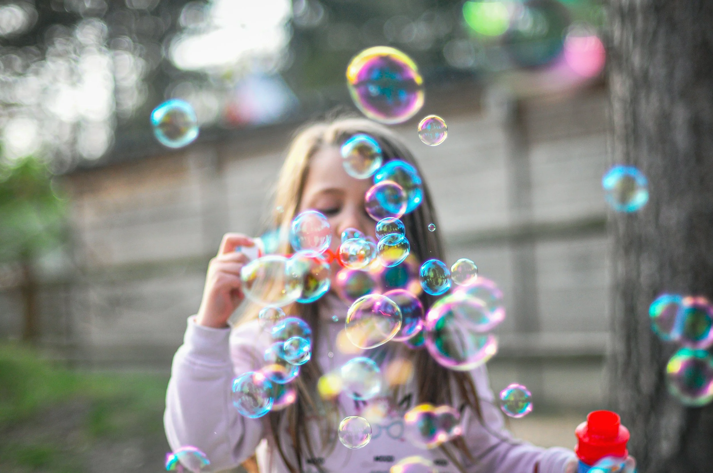 A girl playing with bubbles outdoors in a park or backyard, with a blurred background of trees and a building.