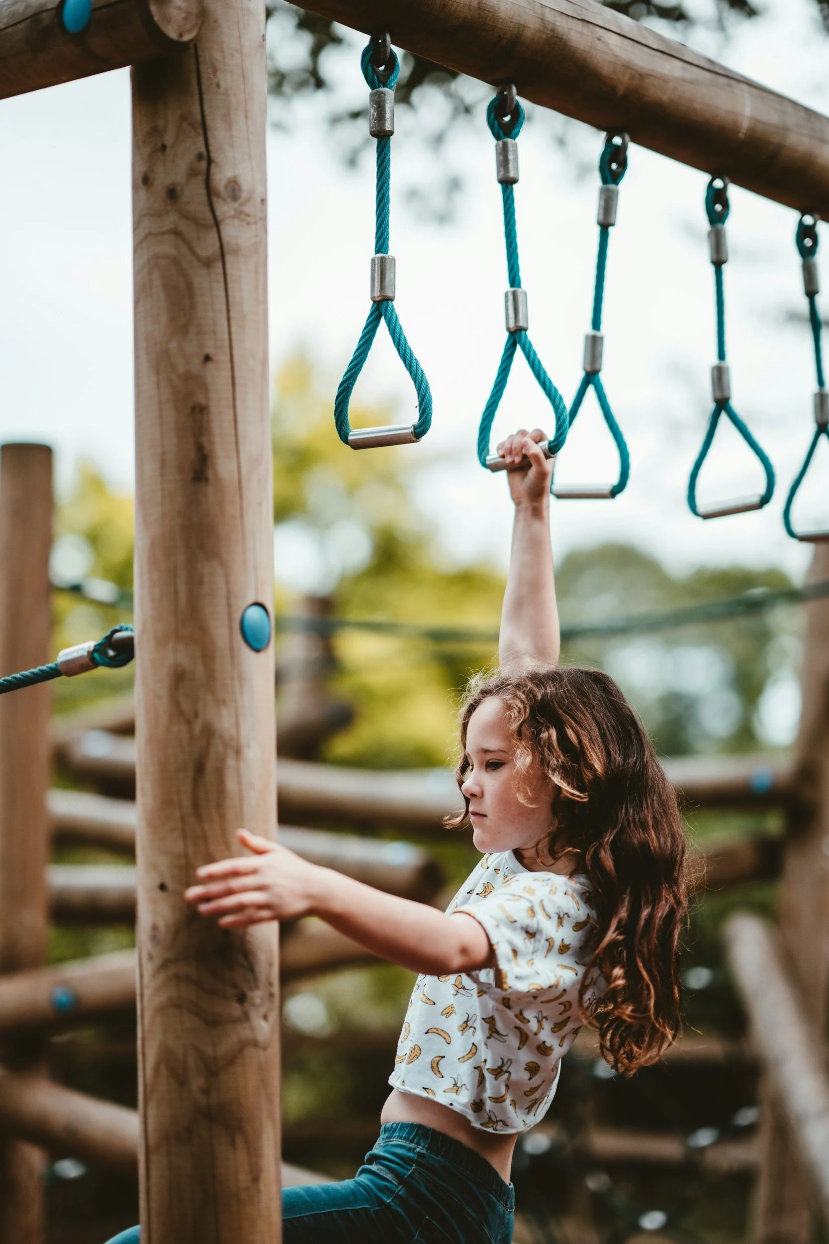 A young girl with long curly brown hair wearing a light-colored shirt with banana prints and blue jeans, enaggeing in movement based play on a wooden playground structure with hanging blue ropes and grips.