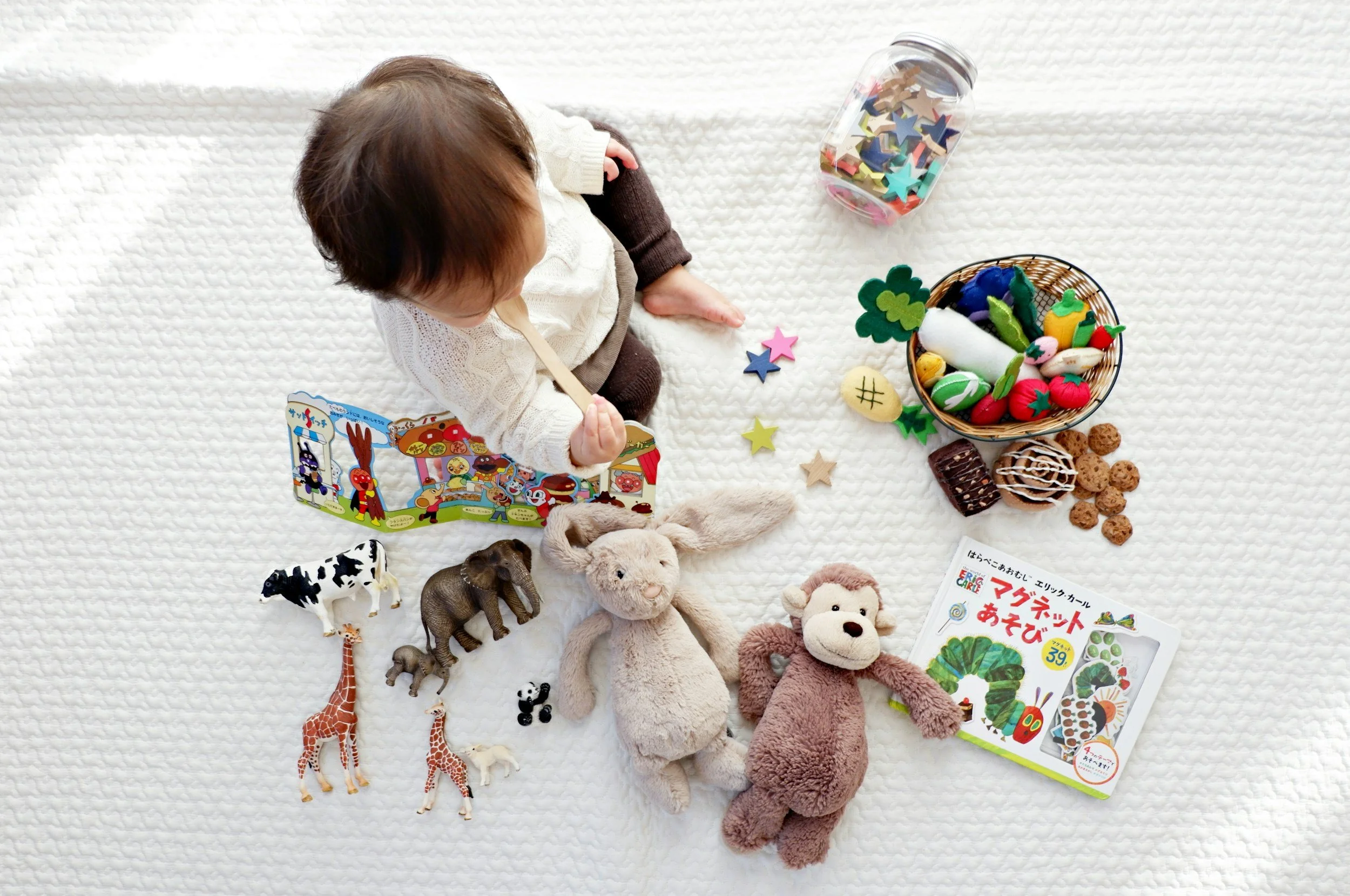 A child sitting on a white textured blanket surrounded by plush toys, toy animals, and a game book, with a jar of colorful foam stars and a basket of felt vegetables and fruits nearby.