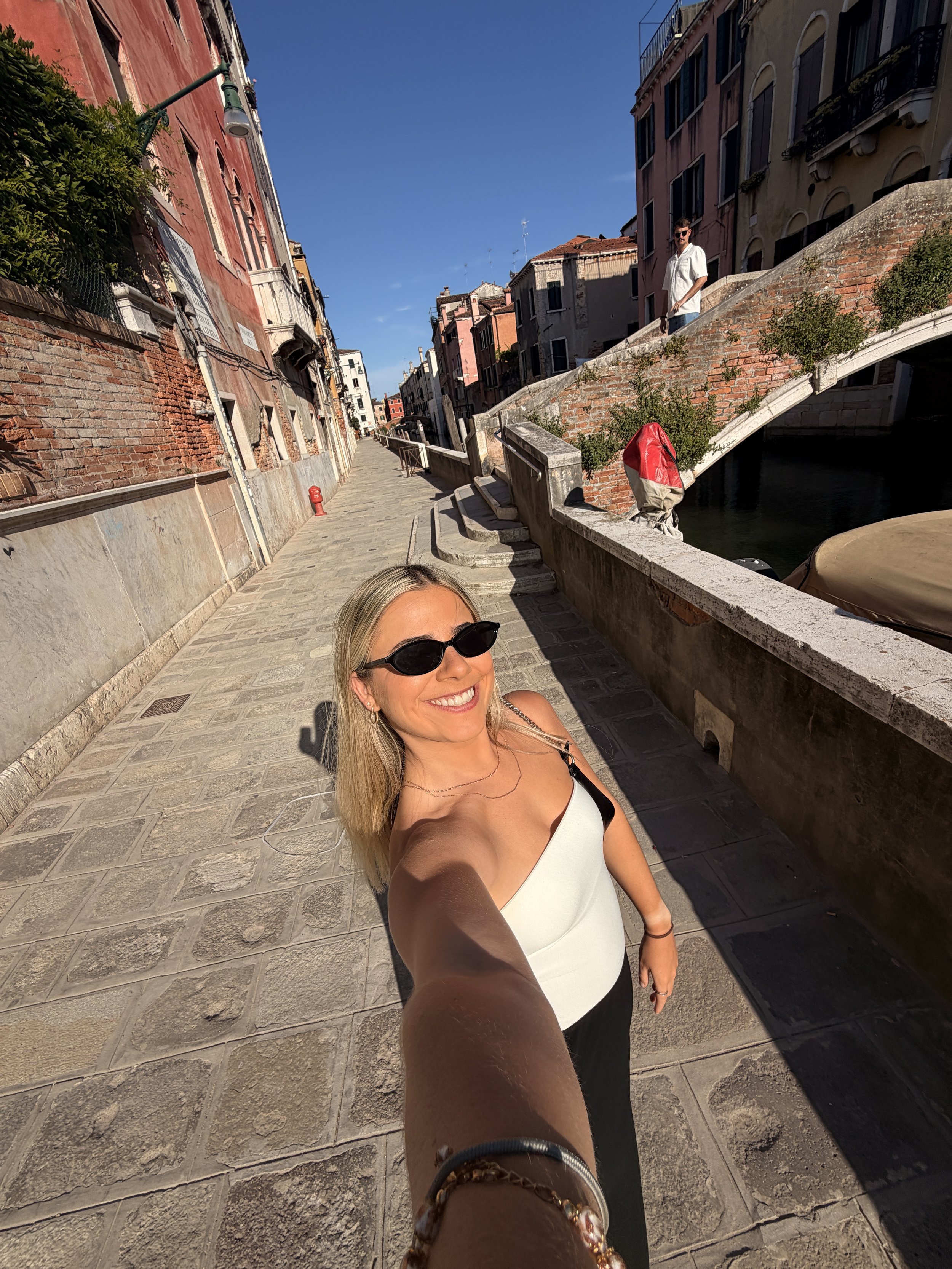 A woman with blonde hair and sunglasses taking a selfie on a sunny day in a European city. Behind her, a canal with a small arched bridge and colorful buildings with traditional architecture line the street.