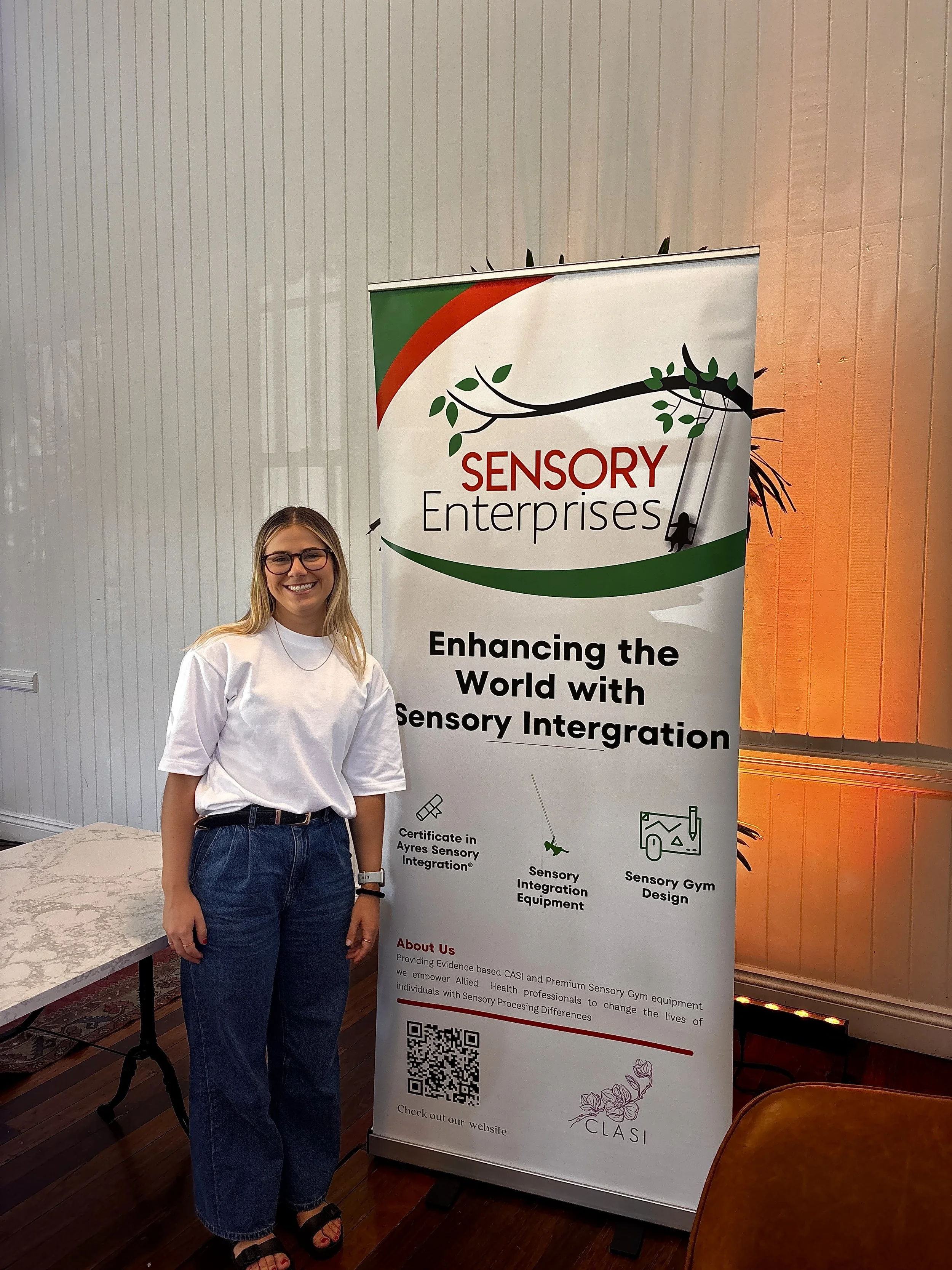 A woman with blonde hair and glasses standing next to a banner for Sensory Enterprises, which promotes sensory integration and related services and qualifications.