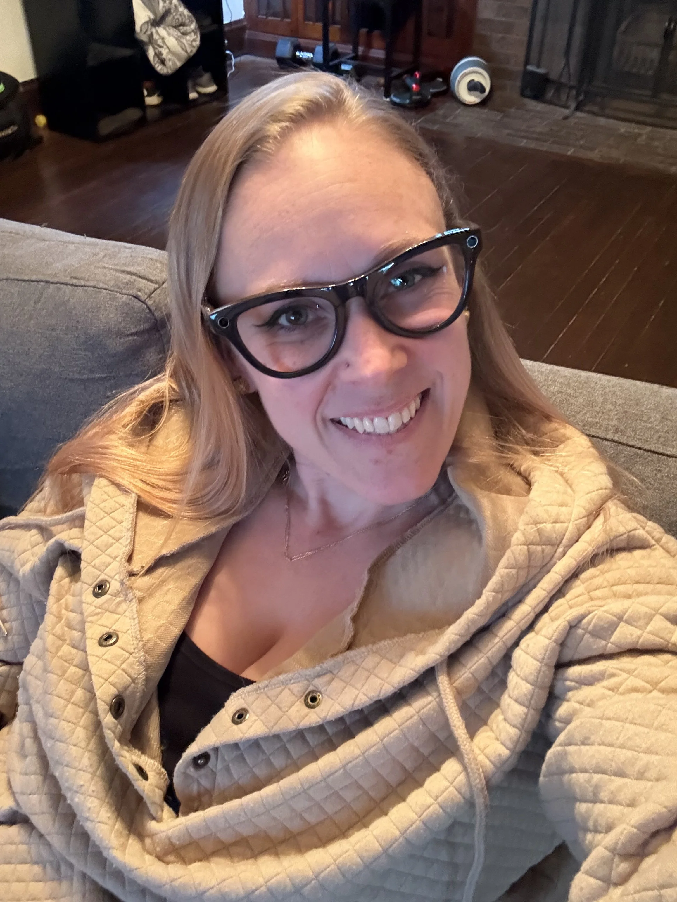 A woman smiling while sitting on a gray couch, wearing black glasses and a beige quilted jacket, with a living room background including a fireplace, wooden cabinet, and various electronic items.