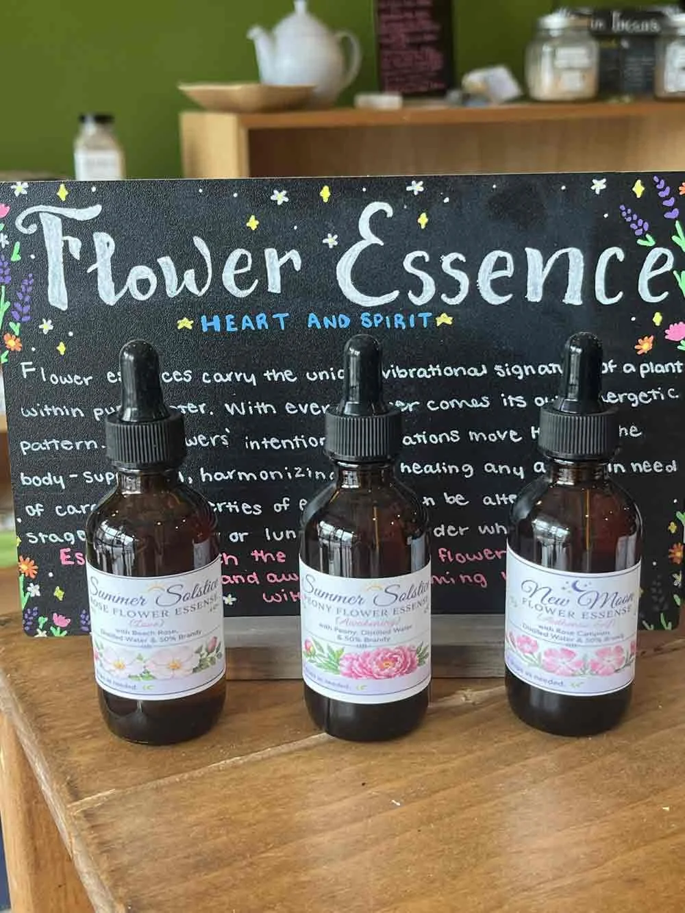 Three bottles of flower essence spray on a wooden surface, with a decorative blackboard in the background labeled 'Flower Essence Heart and Spirit.'
