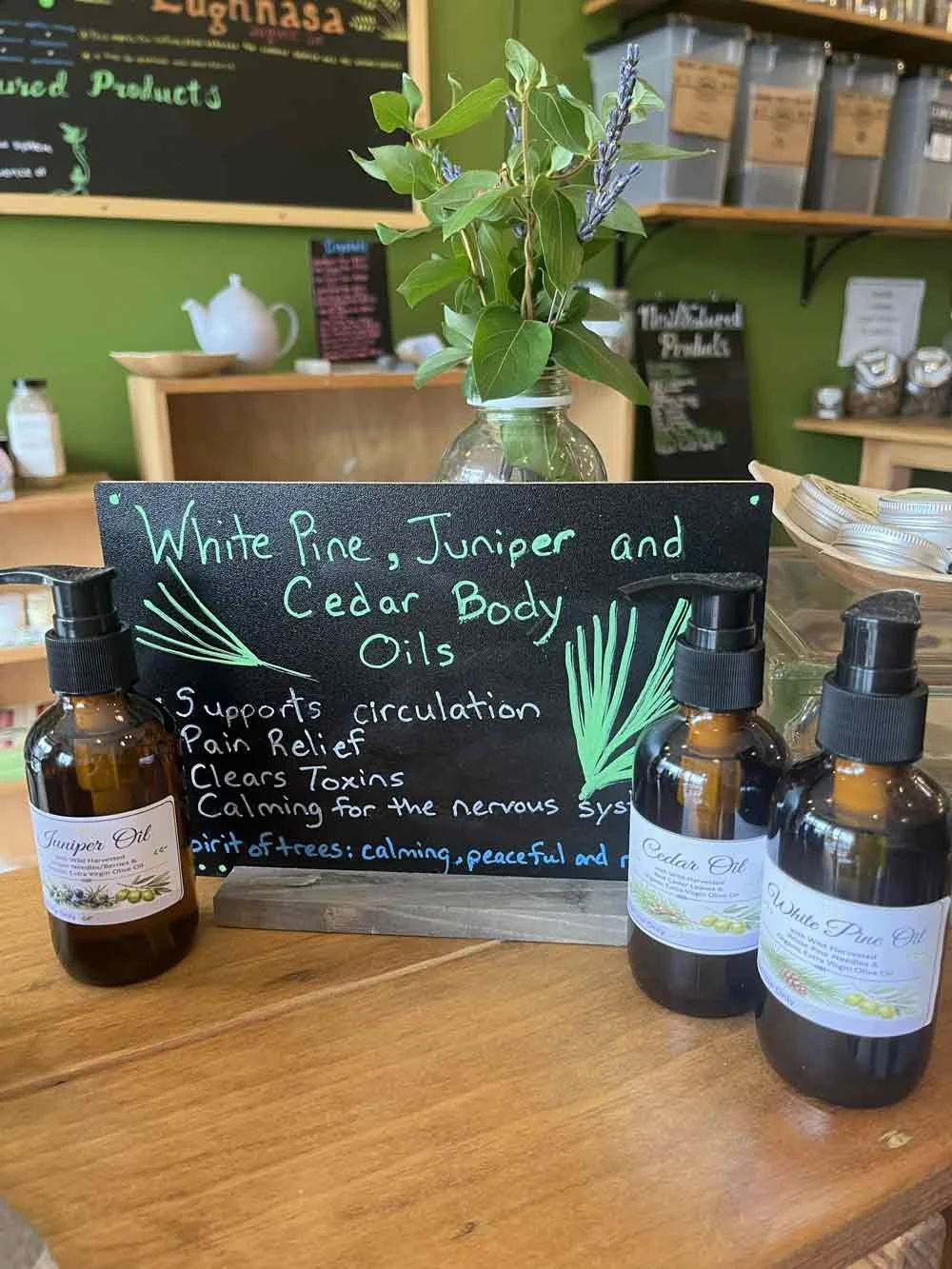 A display of essential oils, including white pine, juniper, and cedar body oils, with a blackboard sign listing their benefits such as supporting circulation, pain relief, clearing toxins, and calming the nervous system. There is a vase with green leaves and lavender, and the background shows a store interior with shelves and a chalkboard menu.