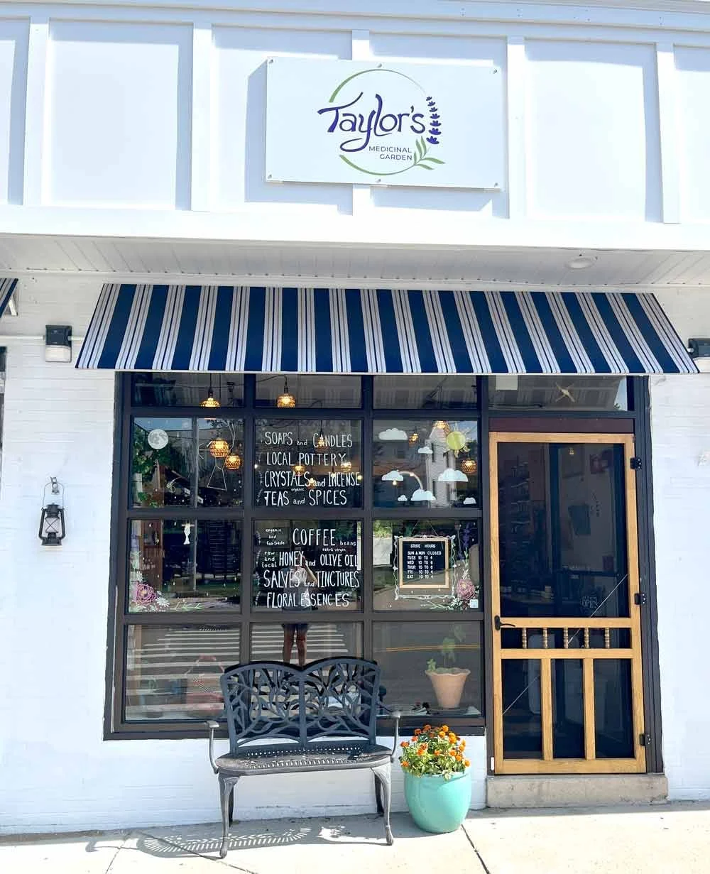 Front of Taylor's Medicinal Garden shop with a striped blue and white awning, black bench, potted flowers, and signage listing products like soaps, candles, pottery, crystals, incense, teas, spices, coffee, honey, and floral scents.