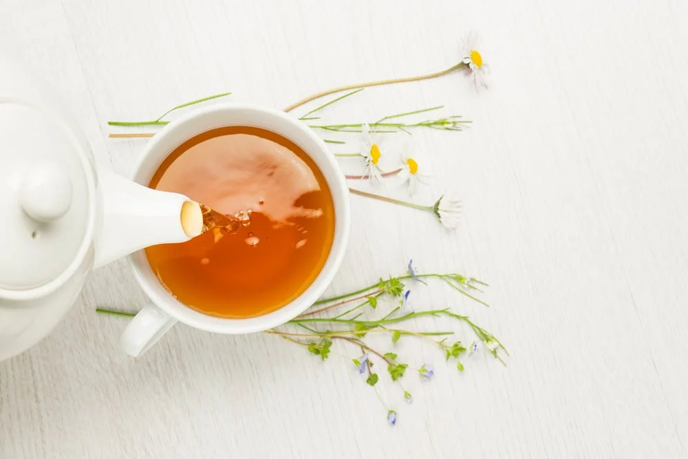 Tea being poured into a white mug surrounded by wildflowers on a white surface.