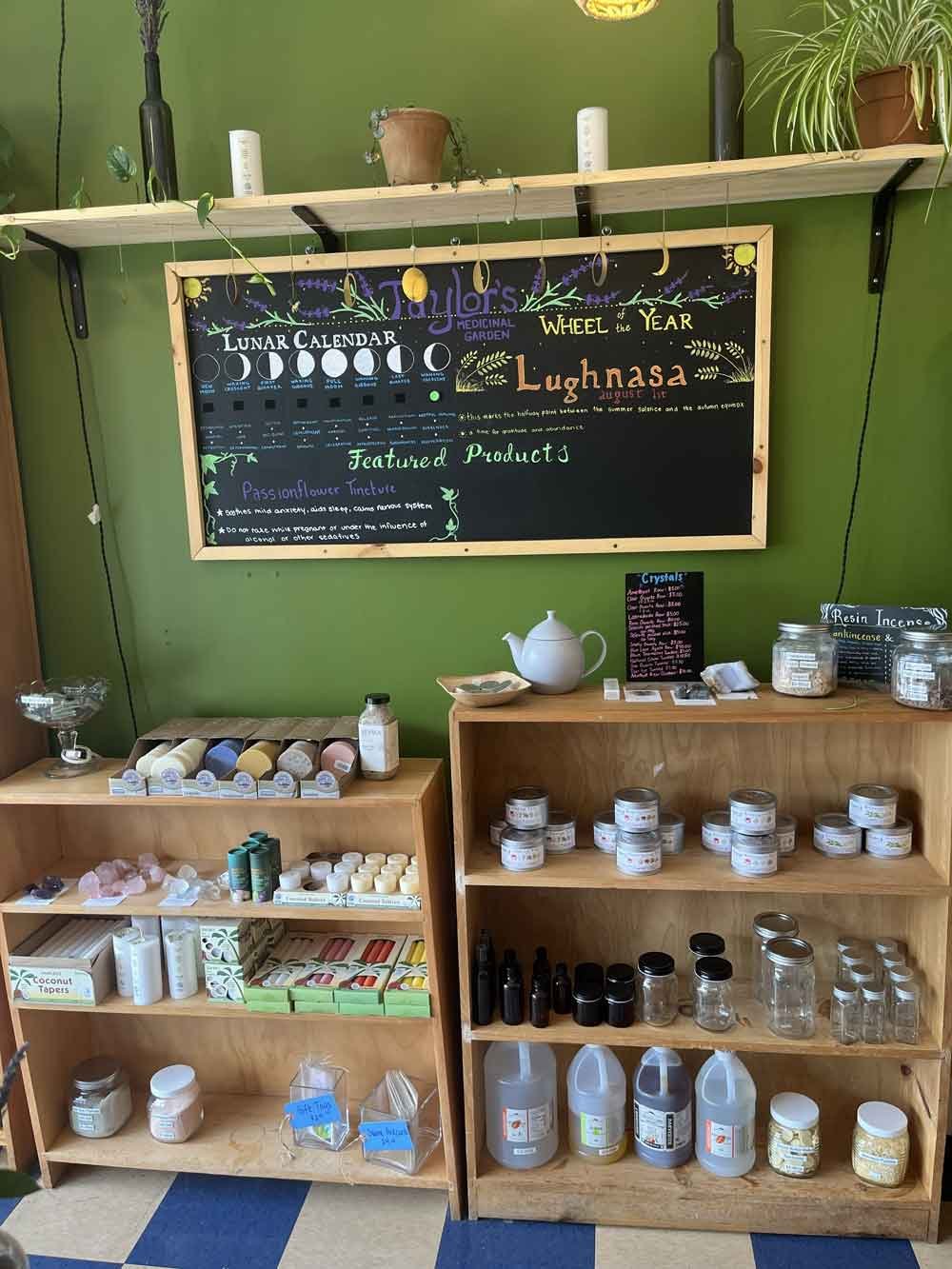 Display of herbs, supplies, and products at a wellness shop with a chalkboard sign titled 'Lunar Calendar' and 'Wheel of the Year Lughnasa' in a green-walled room.