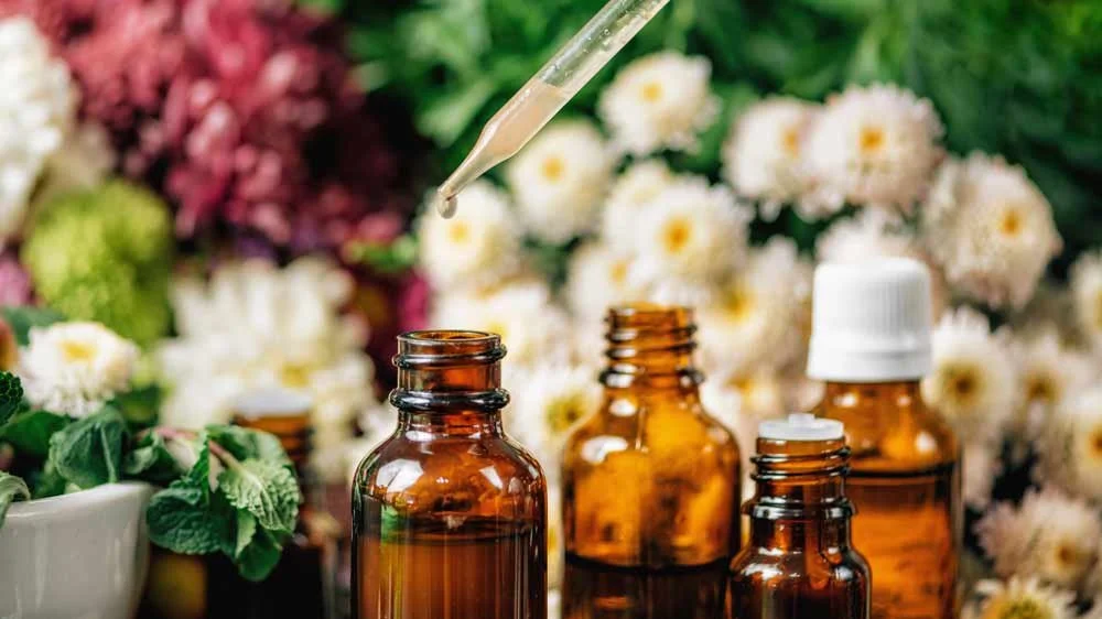 Several brown glass bottles with dropper and white caps on a surface, surrounded by flowers and foliage, with a dropper above releasing a liquid into one bottle.