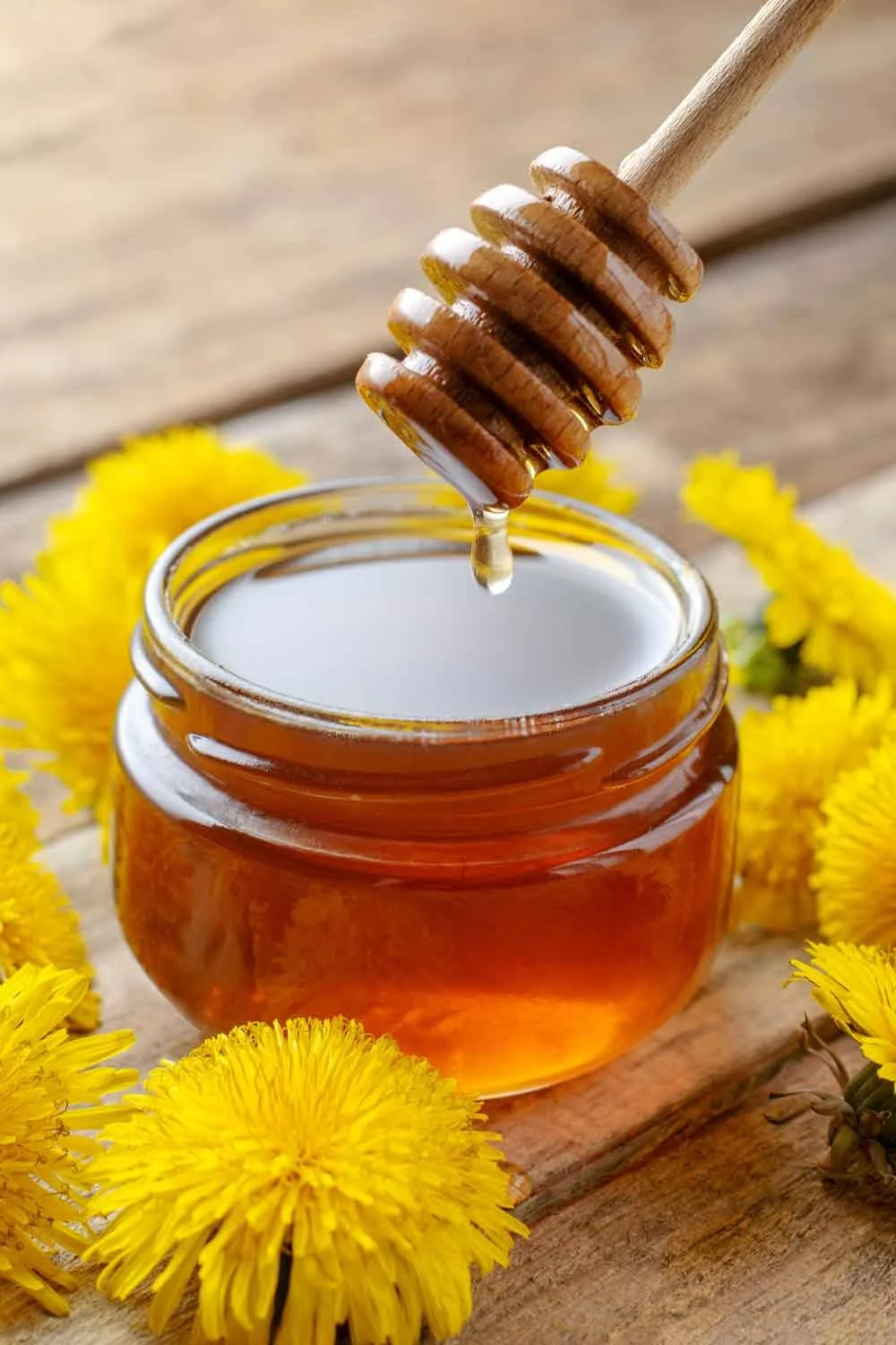Close-up of a jar of honey with a honey dipper dripping honey, surrounded by yellow dandelion flowers on a wooden surface.