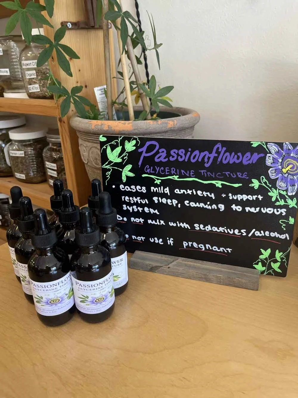 Bottles of passionflower glycerine tincture with a blackboard sign detailing its benefits, including easing anxiety and supporting restful sleep, displayed on a wooden table with a plant and jars in the background.