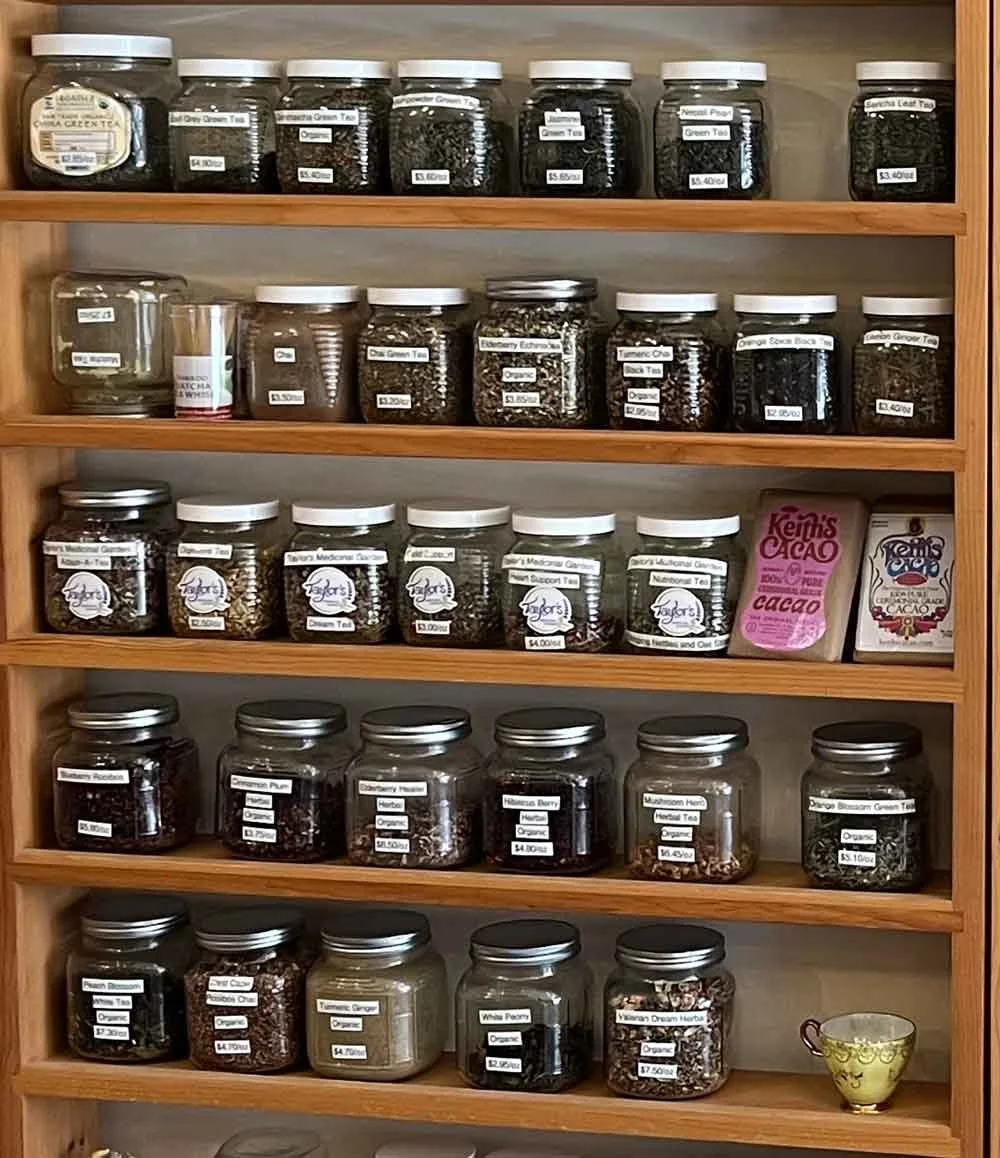 Wooden shelf filled with jars of loose leaf teas, herbs, and medicinal herbs labeled with names and prices, along with tea-related products.