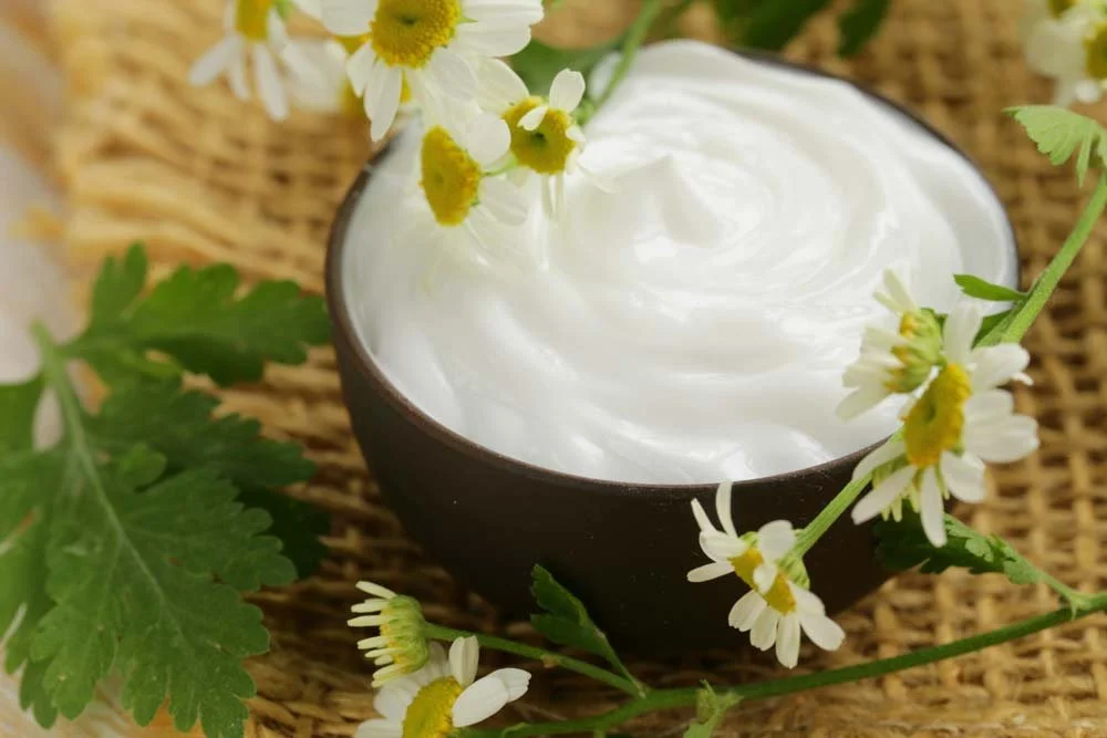 A  container of lotion garnished with chamomile flowers and green leaves, placed on a woven mat.