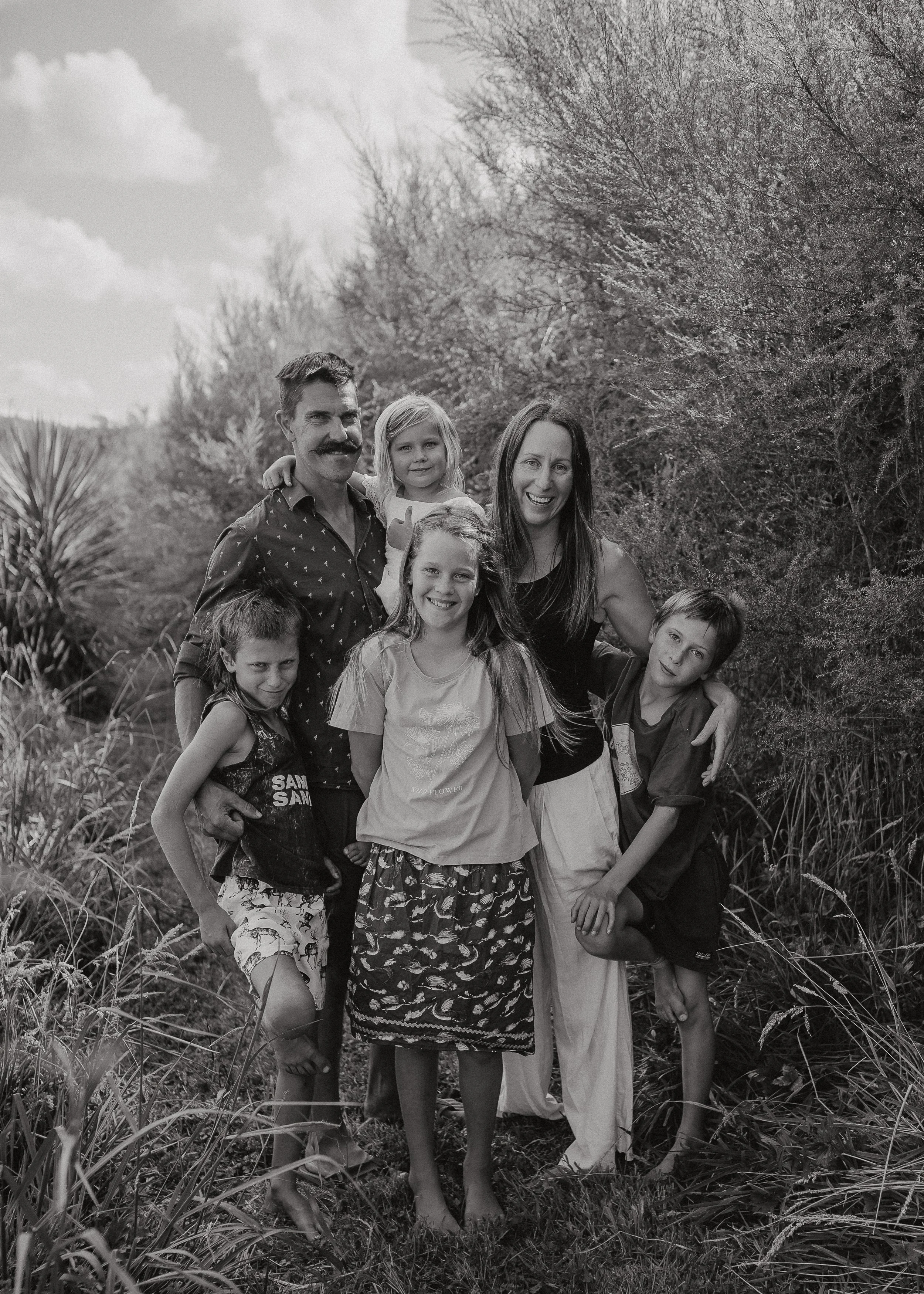 Black-and-white photo of a family of seven standing in a natural outdoor setting with trees and tall grasses, smiling and posing for the camera.