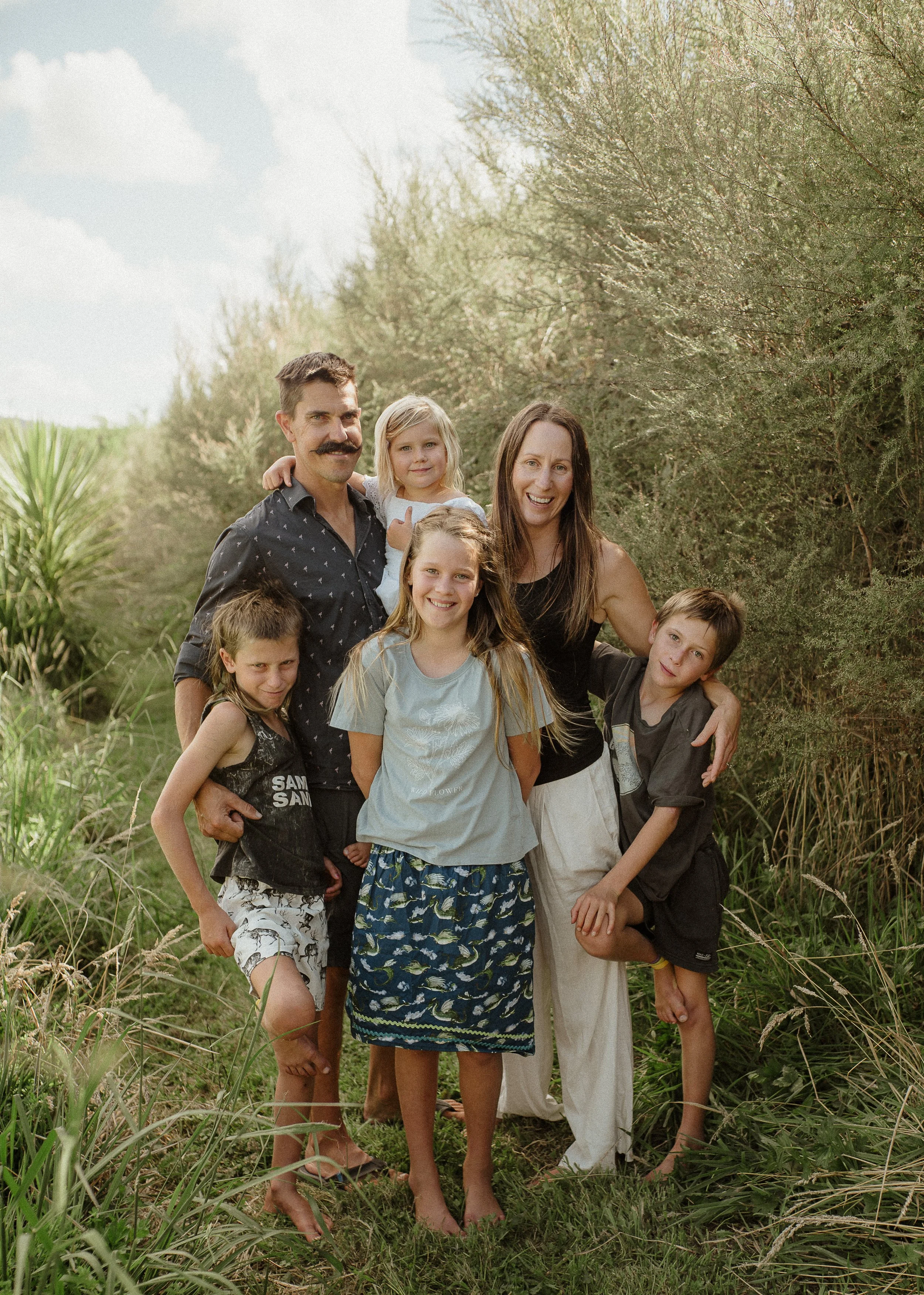 A family of seven smiling and posing outdoors in a natural setting with tall grass and trees, on a sunny day.